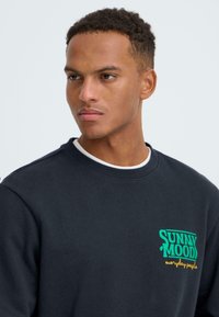 Donkerblauwe sweatshirt met een ronde hals, gestructureerde stof en een kleurrijke voorkant met de tekst "SUNNY MOOD" in groen en geel.