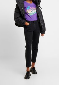 Svart vadderad jacka, lila t-shirt med tropiskt motiv och svarta jeans med hög midja. Svarta snörskor fullbordar outfiten.