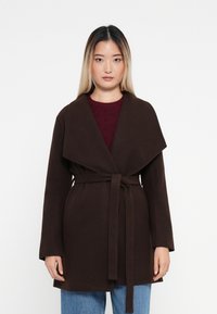 Abrigo envolvente marrón con cinturón y amplio cuello, hecho de una tela texturizada, con mangas largas y bolsillos frontales, superpuesto a una blusa de punto en color burdeos.