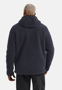Giacca in pile blu navy con finitura morbida e testurizzata, dotata di cappuccio e maniche lunghe. Sul retro presenta un dettaglio di cucitura orizzontale.