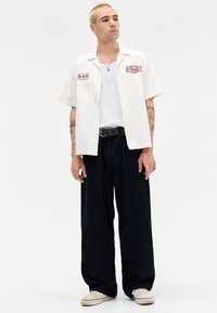 Ung mand med kort blont hår iført en hvid kortærmet skjorte over en hvid tanktop, sorte vide bukser, sneakers og tatoveringer på armene.