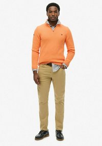Pomarańczowy sweter z dzianiny z zamkiem, khaki spodnie i czarne buty na sznurówki. Strój zawiera jasno niebieską koszulę zapinaną na guziki, noszoną pod spód.
