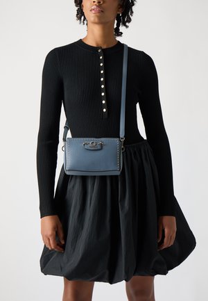 Borsa a tracolla blu in pelle con hardware argentato, dettagli cuciti e tracolla regolabile, indossata sopra una maglietta nera a costine e una gonna voluminosa.
