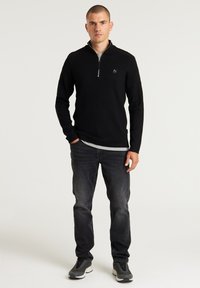 Schwarzer Reißverschluss-Sweater mit langen Ärmeln, aus einem weichen Strickmaterial gefertigt, kombiniert mit dunkelgrauen Jeans und Sneakers. Schmaler Schnitt, lässiges Design.