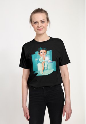 Sort T-shirt med et farverigt grafik af en tegneseriefigur i et badeværelsesmiljø. Designet inkluderer lyseblå fliser og et gulvtæppe.