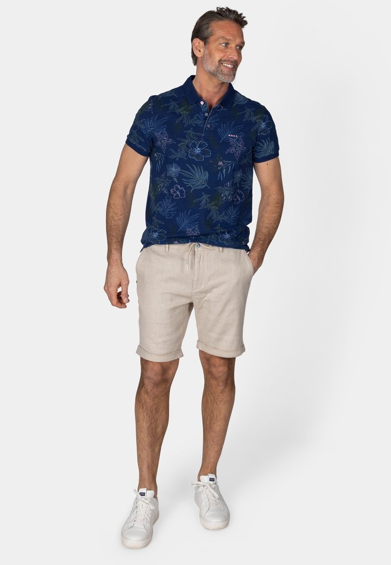 Marineblaues Polohemd mit floralem Muster, kurzen Ärmeln und Knopfleiste mit drei Knöpfen, kombiniert mit hellbeigen Leinen-Shorts mit hochgekrempelten Säumen und weißen Sneakers.