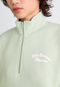 Sweatshirt zippé vert clair en tissu doux. Doté d'un col haut et d'un logo brodé "New Balance Athletics" blanc sur la poitrine.