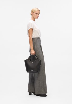 Une femme se tient de côté, portant un haut blanc à manches courtes et un pantalon large gris, tenant un sac à main noir à poignées rondes à ses côtés.