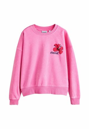 Junge Frau mit lockigem dunklem Haar trägt ein pinkfarbenes Sweatshirt mit roter Hibiskusblume und Coca-Cola-Logo, draußen mit unscharfem Hintergrund.