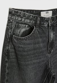 Mustat farkut, joissa on korkeavyötärö, viiden taskun malli ja kontrastinhohtoiset saumat. Niissä on napillinen kiinnitys ja kangas on kevyesti haalistunut.