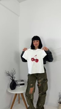 Žena s čiernymi vlasmi drží biely tričko s červeným dizajnom čerešní, oblečená v roztrhnutých olivových cargo nohaviciach, stojí vedľa malej okrúhlej stolíka.
