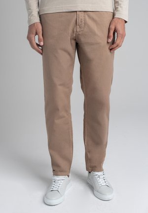 Mężczyzna ubrany w beżowe spodnie, białe trampki oraz kremową koszulę z długim rękawem, stojący na tle jednolitego szarego tła.