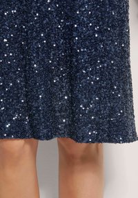 Marineblauwe paillettenjurk met een aansluitende corsage en een uitlopende rok. Het oppervlak is bedekt met kleine, ronde pailletten voor een textuur en reflecterende uitstraling.