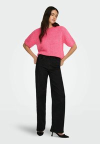 Femme aux longs cheveux noirs portant un pull à manches courtes rose vif, un pantalon large taille haute noir et des talons noirs.