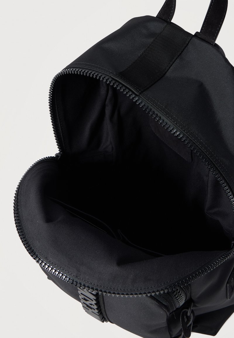 Sac à dos noir avec compartiment principal ouvert montrant l'intérieur vide et la fermeture éclair supérieure partiellement ouverte sur un fond uni.