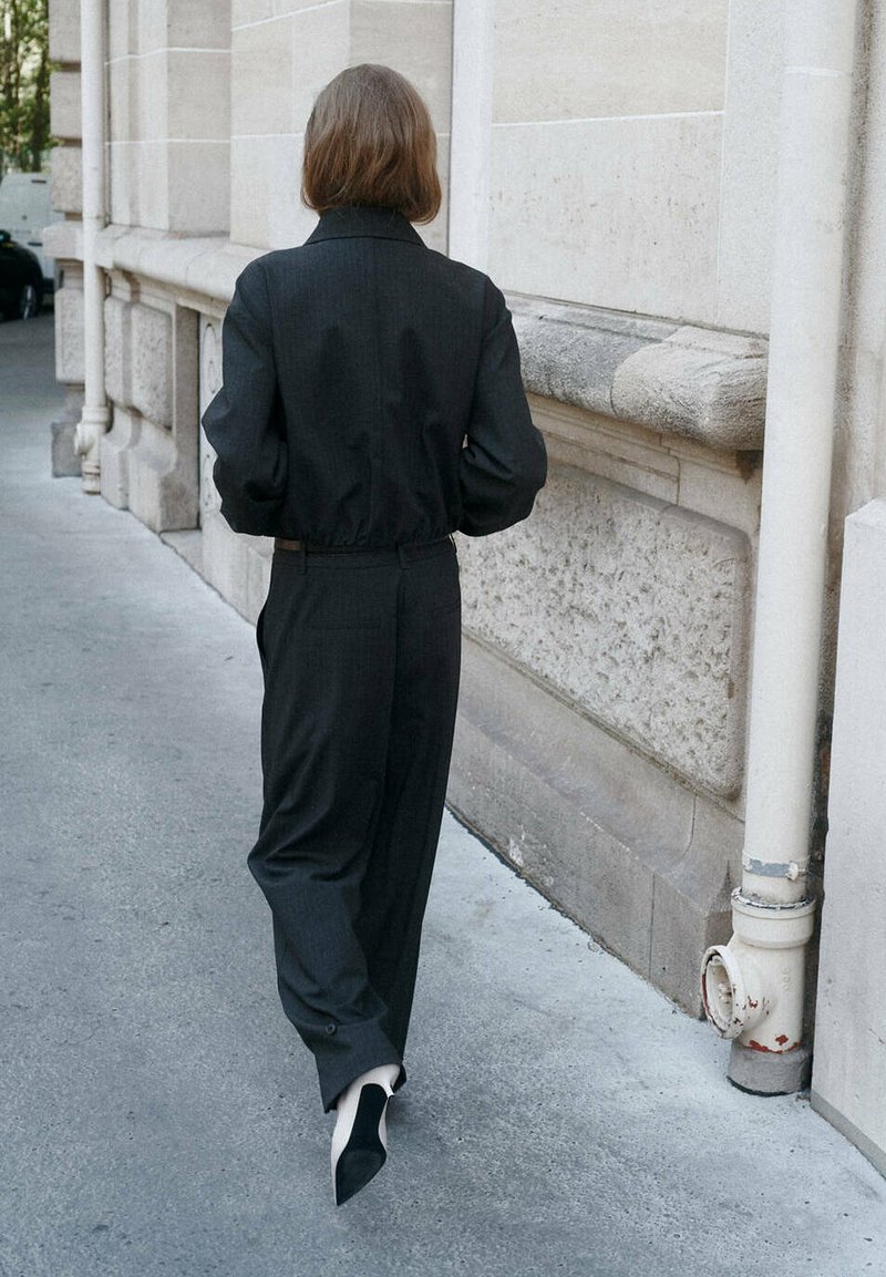 Personne aux cheveux courts et bruns marchant sur un trottoir, portant un costume sombre et des chaussures blanches pointues à côté d’un bâtiment en pierre beige.