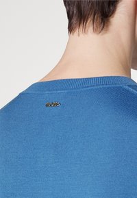 Blå stickad tröja med rundad krage och en liten silverfärgad logotag vid bakhalsen, med en slät textur.