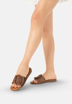 Piernas femeninas que llevan sandalias tipo slide marrones tejidas con grandes hebillas decorativas, de pie sobre un fondo liso gris claro.