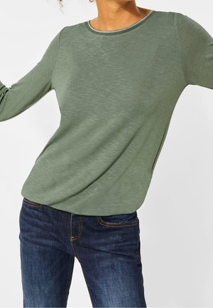 Langärmliges grünes Oberteil aus leichtem Stoff, mit rundem Halsausschnitt und subtiler Textur, kombiniert mit dunkelblauen Jeans.