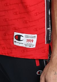 Rød Champion t-shirt med gentaget logomønster. Har et mærke med tekst, herunder "1919" i rød og en sort grafisk detalje. Struktureret stof.