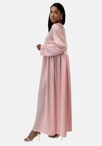 Robe longue en satin rose, coupe ample, manches longues et jupe froncée. Le tissu a une texture brillante, complété par des talons blancs simples.