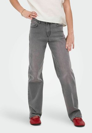 Femeie purtând blugi gri cu picior drept, pantofi roșii fără toc, o cămașă albă cu mâneci scurte și brățări la încheietura mâinii și pe degete, pe partea stângă.