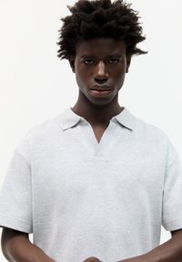 Lys grå polo-shirt med et textured, blødt stof; har en klapkrave og korte ærmer, båret af en person med mørk hud og krøllet hår.