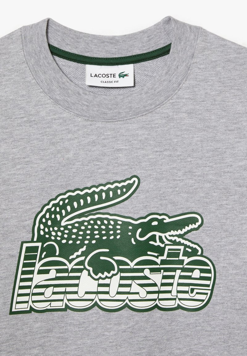 T-shirt in cotone grigio con una grande grafica di un coccodrillo verde e la parola "Lacoste" in lettere a strisce verdi e bianche. Design a vestibilità classica.