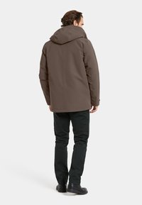 Ruskea vedenpitävä takki hupulla, sileäpintainen ja rento istuvuus. Yhdistetty tummanvihreisiin pantseihin ja mustiin saappaisiin.