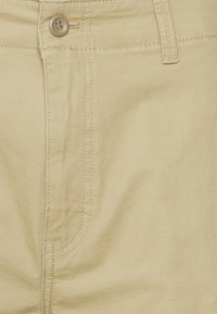 Pantalon en coton beige avec une poche avant, des coutures renforcées et une fermeture à bouton unique. Texture lisse sans motifs visibles.