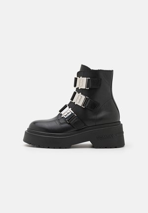 Botte en cuir noire mi-mollet avec une semelle épaisse, trois sangles à boucles argentées et "TOMMY" embossé sur le côté de la semelle.
