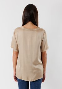 Blusa beige a maniche corte con scollatura rotonda e una texture liscia. Presenta un orlo curvo e una vestibilità rilassata, abbinata a jeans scuri.