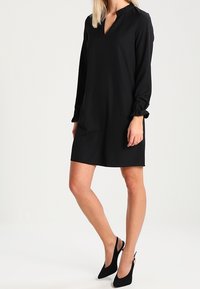 Schwarzes, langärmliges Kleid mit V-Ausschnitt und gerafften Ärmeln, aus glattem Stoff gefertigt. Kombiniert mit schwarzen High Heels.