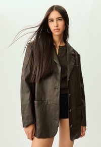 Blazer in ecopelle marrone con vestibilità ampia, due tasche frontali, reverse e chiusura a bottoni. Indossato sopra un top scuro e pantaloni corti.