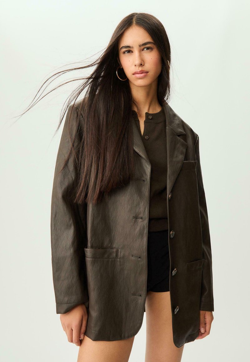 Blazer in ecopelle marrone con vestibilità ampia, due tasche frontali, reverse e chiusura a bottoni. Indossato sopra un top scuro e pantaloni corti.