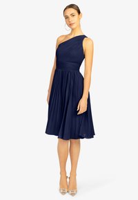 Marineblaues Ein-Schulter-Kleid mit gefaltetem Design, tailliertem Oberteil und schwingendem Rock; kombiniert mit Schuhen mit transparenten Absätzen und großen Creolen.