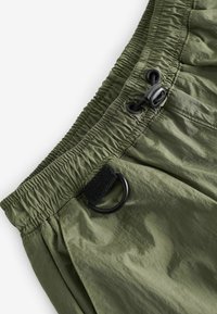 Pantaloni corti verde oliva con cintura elastica, chiusura con cordino e un anello di tessuto nero. Texture liscia, materiale leggero.