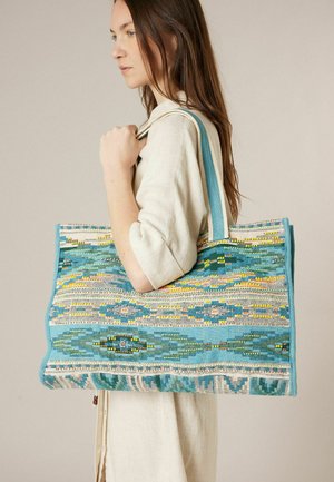 Vrouw met lang bruin haar draagt een grote rechthoekige draagtas met blauwe, gele en witte geweven geometrische patronen over haar schouder.