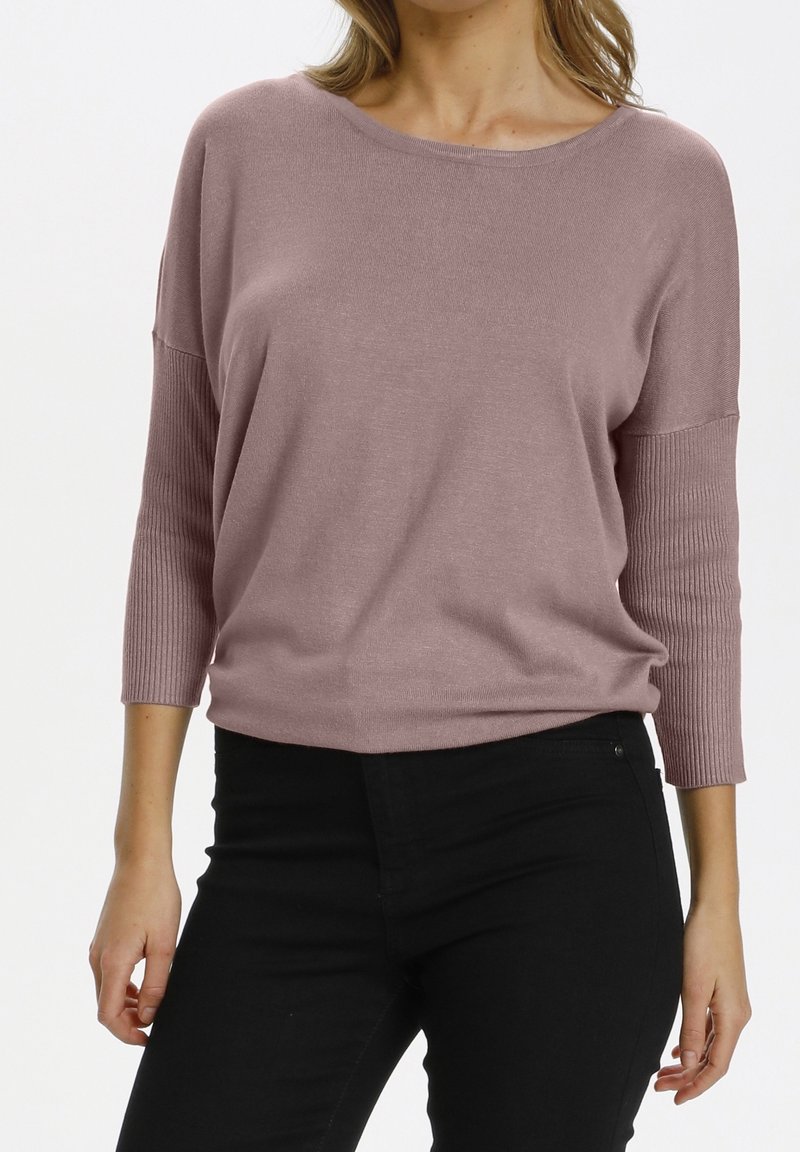 Femme portant un pull mauve à encolure bateau avec des manches 3/4 côtelées et un pantalon noir taille haute sur un fond blanc uni.