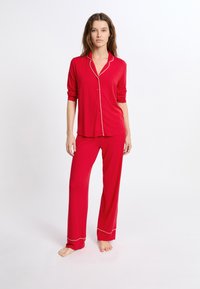 Czerwony zestaw piżamowy z guzikowym topem z białym wykończeniem, długimi rękawami oraz dopasowanymi spodniami o luźnym kroju z białymi lamówkami przy kostkach.