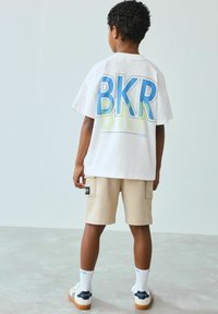 Hvid bomulds t-shirt med stort blåt grafisk "BKR" og grønne detaljer, parret med beige shorts og hvide sokker med blå bogstaver.