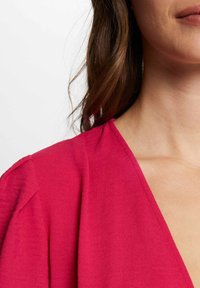 Heldere fuchsia top met een diepe V-hals, textuurstof en uitgesproken schoudernaden, die een strakke, moderne uitstraling toont.