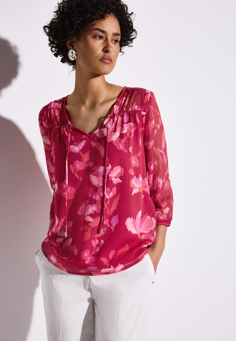 Blusa floreale nei toni del rosa con scollatura a V, maniche a tre quarti e dettagli arricciati sulle spalle. Tessuto leggero e trasparente.