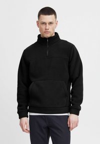 Svart fleece-pullover med høy krage, halvdels glidelås og lomme foran. Har en jevn tekstur og ribbede mansjetter. Hengelapp synlig.
