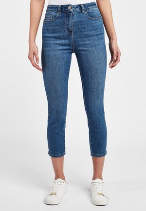 Femme portant un jean skinny bleu court et des baskets blanches, debout devant un fond uni.