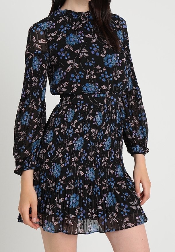 Femme portant une robe noire à manches longues avec des motifs floraux et de baies bleues, une taille cintrée et une jupe plissée.