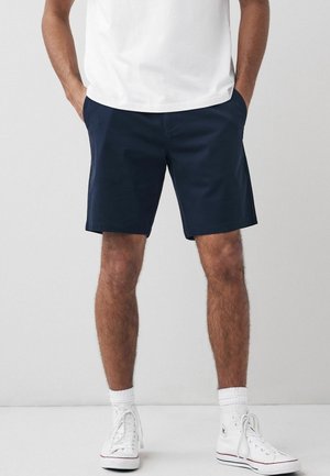 Shorts navy en tissu lisse, avec une coupe droite et des poches latérales, accompagnés de baskets montantes blanches et de chaussettes à hauteur de cheville.