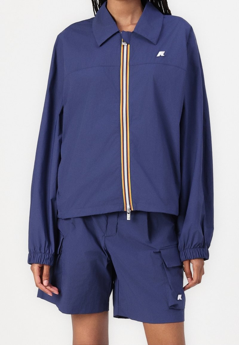 Personne portant une veste coupe-vent bleu marine avec une fermeture éclair rayée et un short cargo assorti avec poche latérale et écussons du logo.
