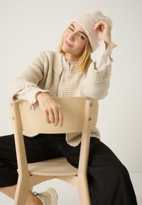 Gilet en tricot beige porté sur une blouse claire à manches à volants, associé à un pantalon noir et des baskets beiges, assis sur une chaise en bois.