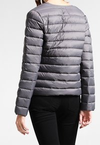 Grå polstret jakke med vandrette sømme, rund halsudskæring, letvægtsmateriale og lange ærmer. Båret over sorte bukser.